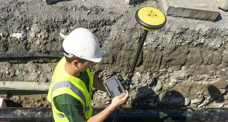 Construction worker in yellow safety vest and helmet in a drench with pipes records points with a Leica iCON CC70 field controller and a Leica iCON GPS 30.